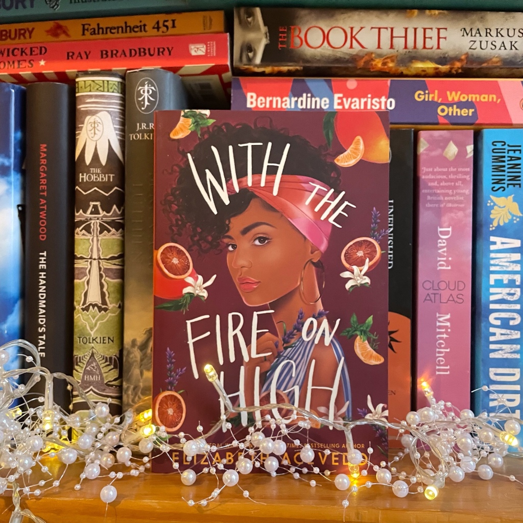 'With the fire on high' by Elizaveth Acevedo stood upright on a bookshelf, fairylights are laid on the shelf. Overlayed on the image is a red box with the word 'reviews'